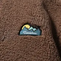 Флісова куртка кавового та бежевого кольорів із високим коміром і нашивкою на грудях.